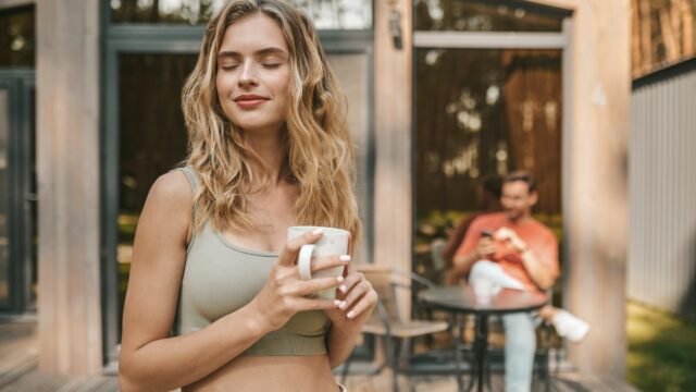 Pretty long-haired blonde woman with a coffee cup standing outsie the cottage